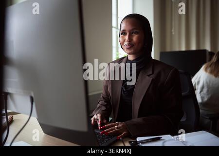 Smiling female tech expert using laptop and computer while sitting at desk in office Stock Photo