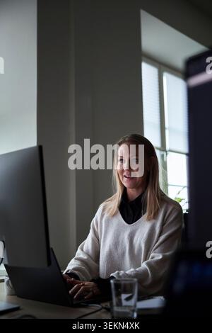 Smiling female business expert using laptop with computer at desk in tech office Stock Photo