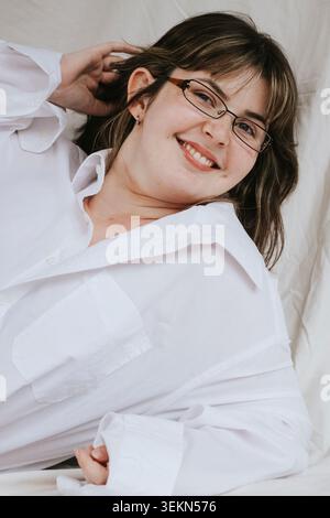 Smiling young woman wearing eyeglasses leaning down against white backdrop Stock Photo