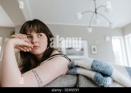Thoughtful young woman leaning on sofa in living room at home Stock Photo