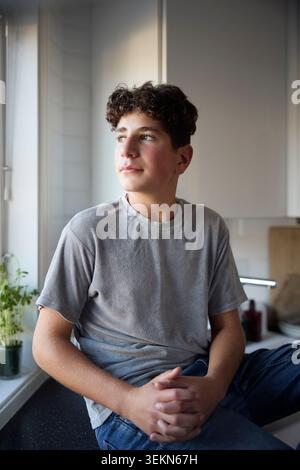Thoughtful boy looking out of window while sitting on kitchen counter at home Stock Photo