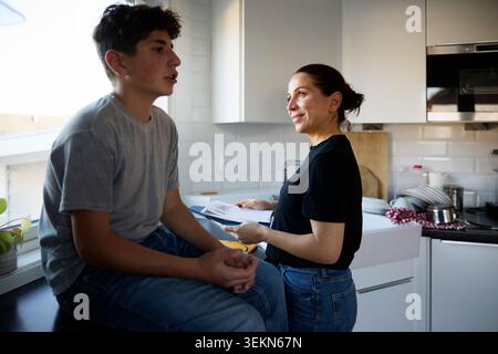 Smiling woman doing dishes and talking with her son sitting on counter in kitchen at home Stock Photo