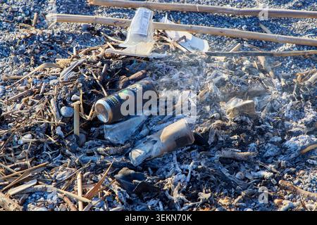 Discarded aluminum cans and plastic trash burnt in a campfire on a pebble beach close up. Stock Photo