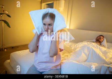 Irritated wife sitting on bed covering ears with pillow to block snore sound while man sleeping Stock Photo