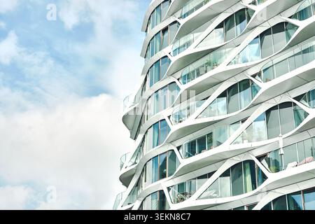 Contemporary multi-story tower with flowing curved balconies and glass windows, viewed from below against a bright blue sky with soft clouds Stock Photo