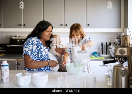 Smiling girl using electric beater with assistance of mother standing beside with toddler in kitchen Stock Photo