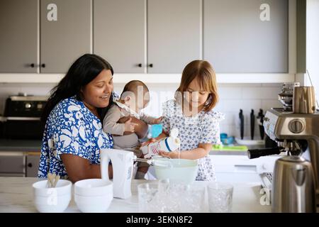 Mother carrying toddler son while assisting daughter pouring cream in bowl at kitchen island Stock Photo