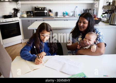 Mother sitting with male toddler while assisting daughter in studies at home Stock Photo