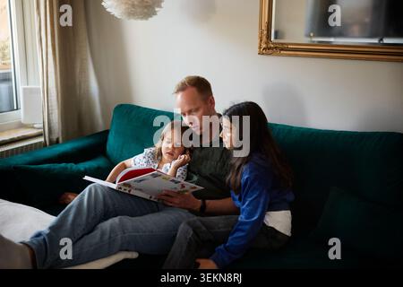High angle view of father reading picture book to daughters while sitting on sofa in living room at home Stock Photo