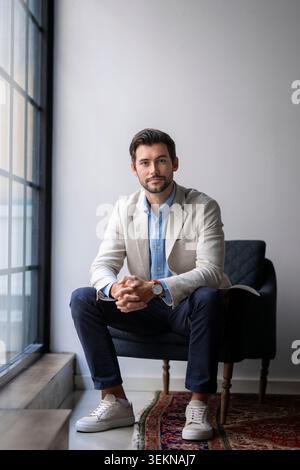 Portrait of young businessman sitting with hands clasped on armchair near window at startup office Stock Photo
