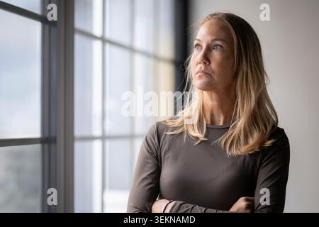 Contemplative blond businesswoman looking away while standing with arms crossed in office Stock Photo