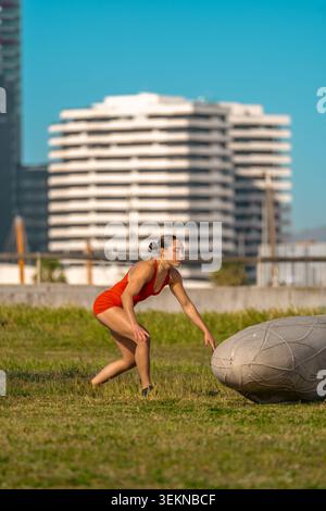 Side view of a young athletic woman in a red suit landing on the grass after performing a backflip or gymnastic acrobatics in a city park. Stock Photo