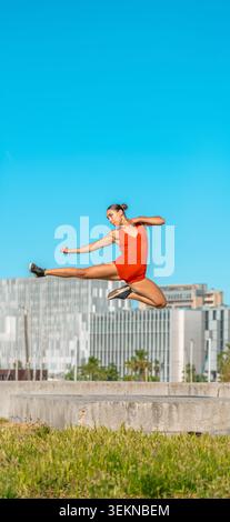 Athletic woman in red bodysuit performing dynamic mid-air jump Flying Side Kick outdoors against city skyline and blue sky. Stock Photo