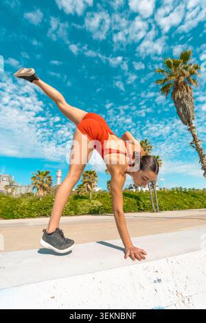 Low angle view of a strong young woman doing a one-handed cartwheel or handstand variation on a stone ledge at a coastal promenade Stock Photo
