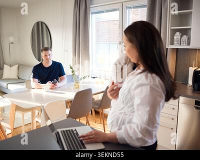 Woman multi-tasking while talking with man sitting at dining table in home Stock Photo