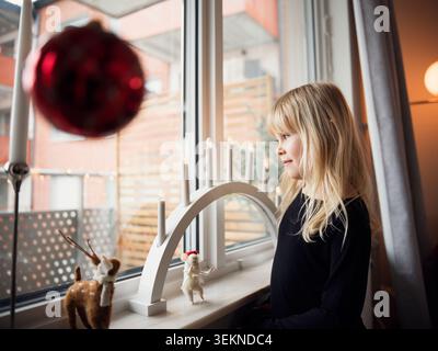 Contemplative blond girl looking out through window while standing near figurines and electric candles at home Stock Photo