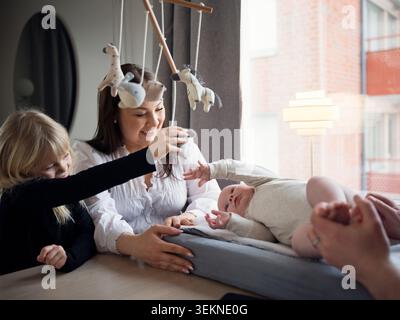 Smiling mother looking at daughter holding toy while playing with baby brother lying down at home Stock Photo
