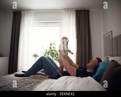 Happy father playing with toddler son while lying on bed in bedroom at home Stock Photo