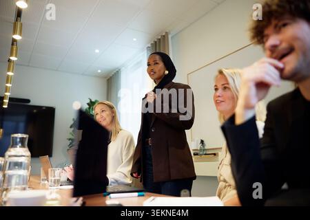 Businesswoman sharing ideas while standing amidst coworkers in office meeting Stock Photo