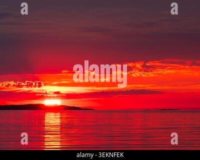 Bright red sun setting over calm sea with dark islands Stock Photo