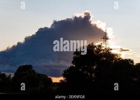 Dramatic summer sunset silhouette featuring a looming storm cloud contrasted by bright sun rays behind a transmission tower and dark foliage Stock Photo