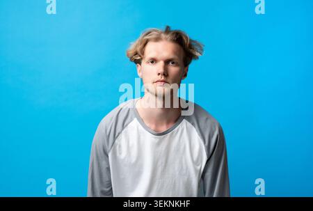 Young man with blond hair and a neutral expression standing directly facing the camera, wearing a casual long-sleeve t-shirt against a striking blue Stock Photo