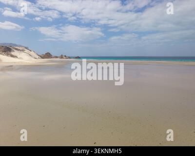 Wide sandy beach with shallow wet shore, turquoise sea, pale dunes and rocky cliffs under a soft cloudy sky. Calm coastal landscape with open space an Stock Photo
