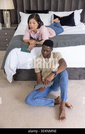 Diverse couple reclining in master bedroom, woman holding green passport, man using tablet. Bed, duo, device, documents, relaxed, interior, lifestyle Stock Photo