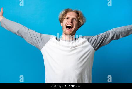 Young man with arms outstretched celebrating success, experiencing joy and freedom, shouting in excitement with an open mouth against a blue Stock Photo