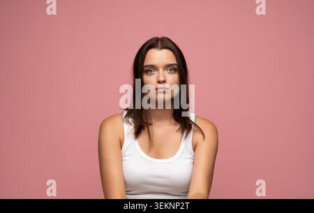 Young woman with serious expression posing for a studio portrait, showing neutral emotion on pink background Stock Photo