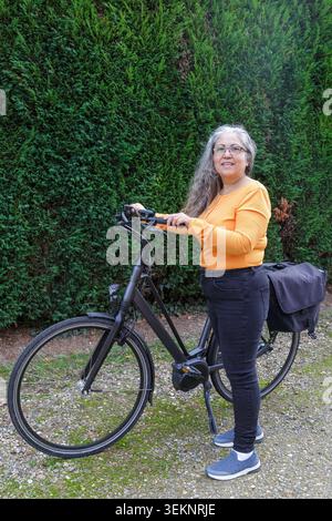 Adult woman standing next to a black bicycle on unpaved rural road, smiling expression on face, dressed in casual clothes, orange shirt and black pant Stock Photo
