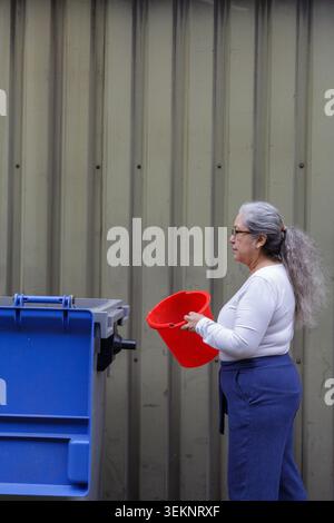 Female worker with a red bucket in hand walks towards industrial waste container to deposit garbage, sheet metal wall in background, waste management Stock Photo