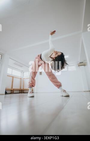 Young female dancer stretching in bright studio wearing pink joggers and white sweater Stock Photo