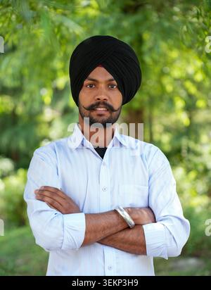 A close-up portrait of a young Punjabi Sikh man wearing a traditional black turban and a light blue shirt, standing confidently with arms crossed. Stock Photo