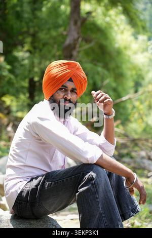 Vertical shot of a young Punjabi Sikh man wearing a bright orange turban and a metal Kada bracelet sits on a rock, looking at the camera. Stock Photo
