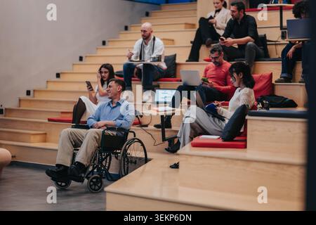 Students in a modern lecture hall with a man in a wheelchair using laptops and phones Stock Photo