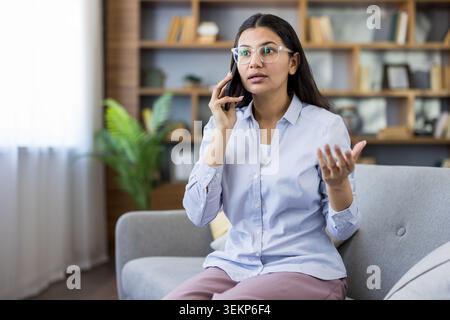 Woman wearing eyeglasses having a serious conversation on her smartphone while sitting on a sofa at home, gesturing with her hand, discussing important matters or feeling frustrated Stock Photo