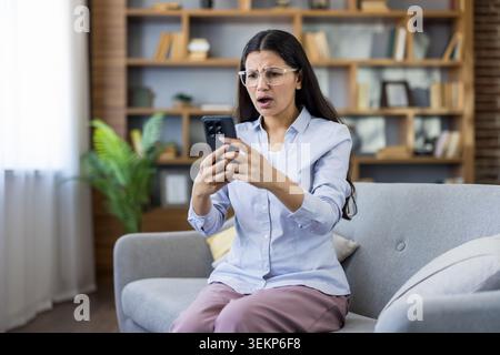 Young woman sitting on sofa at home, upset and frustrated while reading a shocking or disappointing message on her smartphone, reacting to unexpected bad news Stock Photo