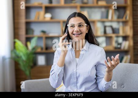 Young woman wearing eyeglasses making a phone call, actively engaging in conversation with an open hand gesture, sitting at home in a modern living room Stock Photo