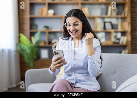 Happy young woman in glasses triumphing and celebrating success, holding a smartphone and expressing excitement while sitting on a couch at home after receiving good news Stock Photo