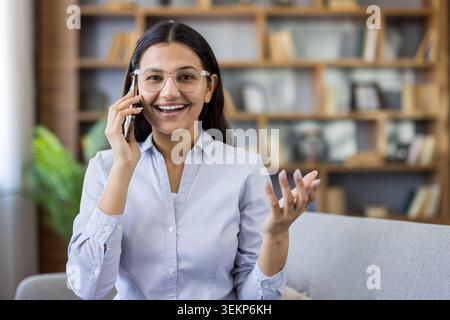Smiling young adult woman wearing glasses and a striped shirt, holding a mobile phone to her ear and gesturing, having a pleasant conversation indoors Stock Photo