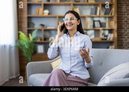 Young woman wearing glasses and a light blue shirt, enjoying a pleasant phone conversation while sitting comfortably on a modern sofa in a bright home office, actively communicating Stock Photo