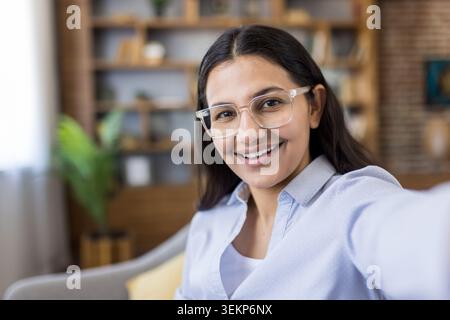 Young happy indian woman with eyeglasses smiling, looking at camera and recording a video or having an online consultation from home, showcasing modern communication Stock Photo