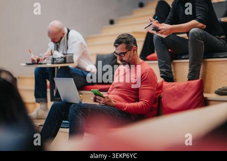 Man using laptop and smartphone while seated in a casual workshop with tiered seating and cushions Stock Photo