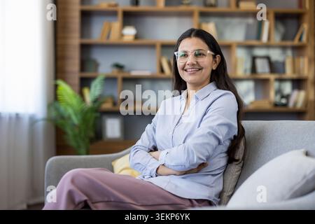Young happy professional woman, wearing eyeglasses and a blue shirt, sitting comfortably on a grey sofa with arms crossed, smiling in a modern living room setting with a bookshelf Stock Photo