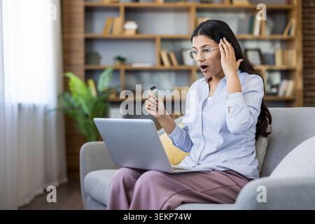 Young woman sitting on a couch at home, looking shocked and stressed while holding a credit card and using a laptop for online shopping, possibly facing financial trouble Stock Photo