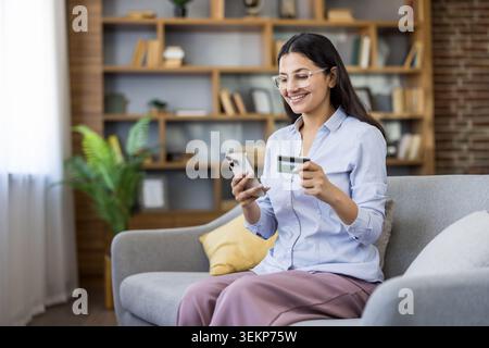 Young smiling woman sitting on a sofa at home, using her smartphone and holding a credit card, enjoying convenient online shopping and secure payment transactions Stock Photo