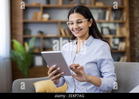 Young woman wearing glasses sitting at home, happily holding and operating a digital tablet, connecting with technology for remote work, education, and digital lifestyle Stock Photo