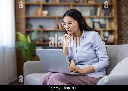 Young woman in glasses sitting on a sofa at home, analyzing information and contemplating solution for a complex problem on her laptop computer, working remotely Stock Photo