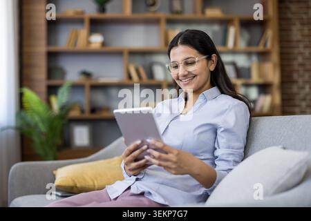 Young indian woman wearing glasses relaxing on a couch at home, smiling while browsing content on a digital tablet, representing modern technology use and comfortable leisure Stock Photo